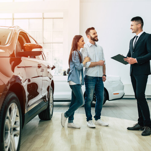 A couple talking to a car salesman in a dealership, with a red SUV in the foreground.