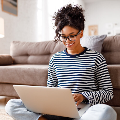 Person with curly hair and glasses uses a laptop on the floor in a cozy living room.