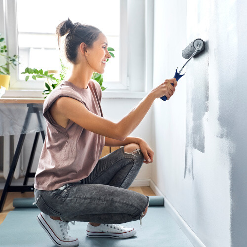 Woman kneeling and painting a wall light gray with a roller.