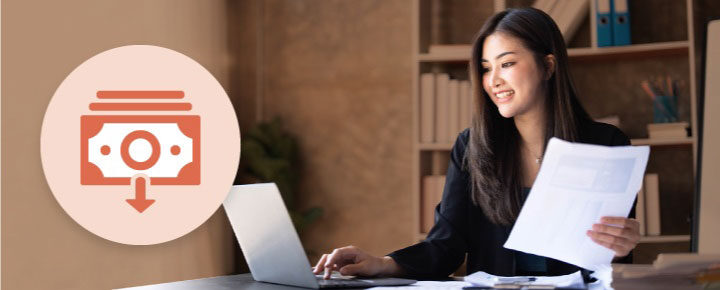 Woman in black blazer working on a laptop while holding a document; icon of a banknote with a downward arrow on the left.
