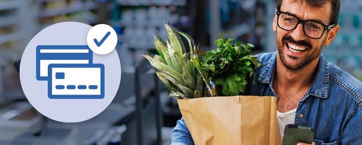 A round icon of a credit card with a checkmark, and a man holding his smart phone and a grocery bag of produce in a store.