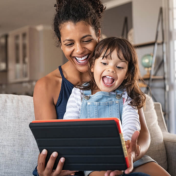 A woman and her daughter, smiling looking at an electronic tablet.